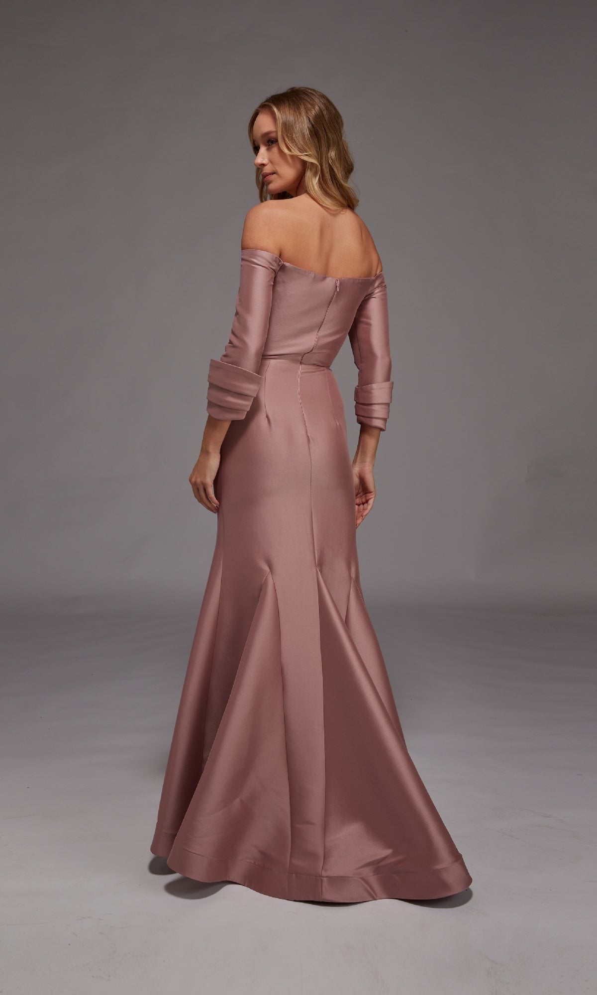 Woman in a mauve Alyce 27782 long formal dress stands facing away, showing the fitted stretch mikado bodice and flared mermaid skirt against a neutral studio background.
